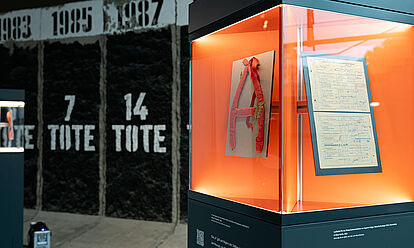 Eine Glasvitrine, in der sich ein aus Alufolie und Krepppapier gebasteltes „A“ sowie ein Dokument befinden.