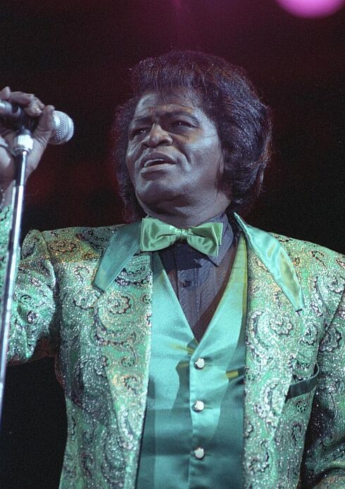 Farbaufnahme von James Brown, der einen bunten Anzug mit grüner Fliege trägt und mit der rechten Hand ein an ein Stativ befestigtes Mikrofon festhält.