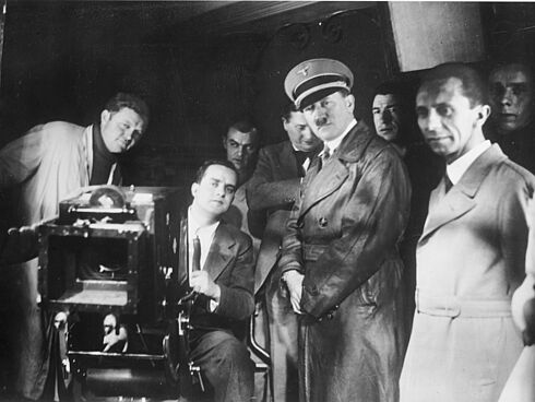Hitler und Goebbels stehen neben einem hinter einer Kamera sitzenden Mann. Dahinter sind weitere Herren zu sehen.