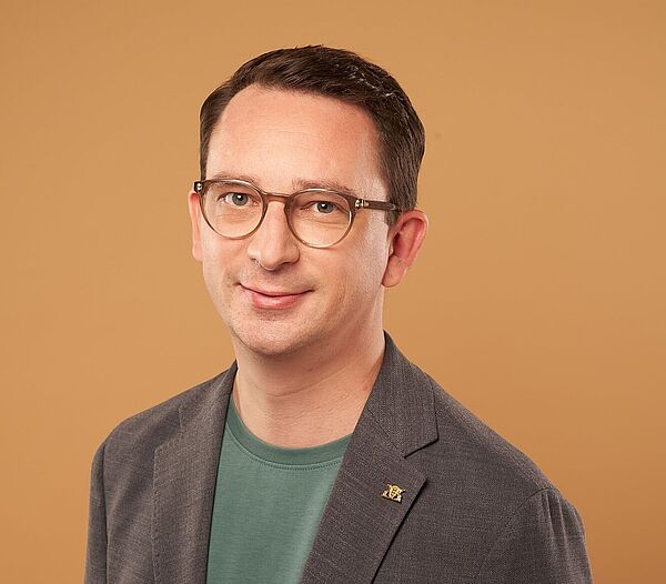 Der Vorsitzende Jonas Weber (MdL) Ein Mann mit kurzen dunklen Haaren in grauem Sakko steht lächelnd vor einer braun-orangenen Wand.