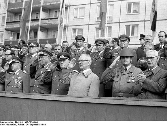 [Auf einer Tribüne stehen in drei Reihen Männer, überwiegend in Uniform, andere im Anzug. Viele salutieren, einige applaudieren. Es ist ein schwarzweißes Foto.]