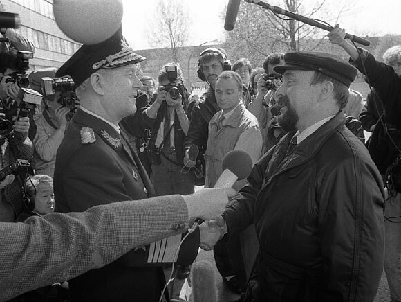 Rainer Eppelmann (r.), Minister für Abrüstung und Verteidigung der DDR, mit seinem Vorgänger, dem Minister für Nationale Verteidigung, Admiral Theodor Hoffmann (l.), bei der Amtsübernahme im April 1990. Eppelmann in Zivil und Hoffmann in Uniform schütteln sich die Hände. Begleitet wird die Übergabe von zahlreichen Reportern mit Mikrofonen und Kameras.