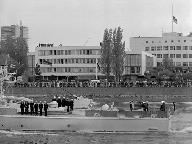 Das Schnellboot „Kondor“ mit dem aufgebahrtem Sarg von Konrad Adenauer passiert das Bundeshaus in Bonn. Die Bundesflaggen wehen auf Halbmast. Eine Menschenmenge beobachtet die Vorbeifahrt vom Rheinufer aus.