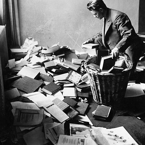 Ein junger Mann kniet vor einem ungeordneten Bücherhaufen und sortiert Bücher in einen großen runden Korb. Das Foto ist in Schwarz-Weiß aufgenommen.