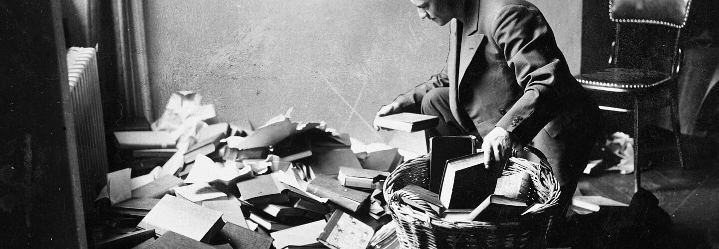 Collection of banned literature, 10 May 1933 Ein junger Mann kniet vor einem ungeordneten Bücherhaufen und sortiert Bücher in einen großen runden Korb. Das Foto ist in Schwarz-Weiß aufgenommen.