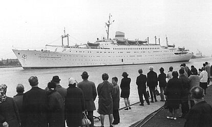 Das DDR-Kreuzfahrtschiff „Völkerfreundschaft“ beim Ablegen im Hafe von Warnemünde. Im Vordergrund stehen Zuschauerinnen und Zuschauer.
