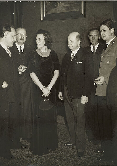 Filmregisseurin Leni Riefenstahl mit u. a. dem Präsidenten der Reichsfilmkammer, Prof. Dr. Oswald Lehnich, bei der Veranstaltung zu „40 Jahre deutsche Kinotechnik“ Leni Riefenstahl steht in Abendgarderobe zwischen mehreren Herren in Anzügen. Sie lächelt in Richtung eines sprechenden Mannes.