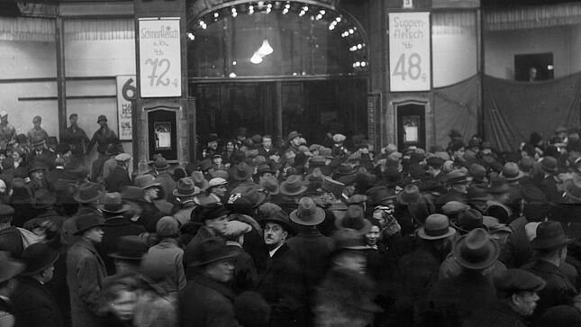 Das Bild zeigt eine Menschenmenge vor dem Warenhaus Tietz. Rechts und Links vom Eingang sind Angebotsschilder für Fleischwaren angebracht. In der Bildmitte schaut ein Man mit Fedora und Schnauzer in die Kamera.