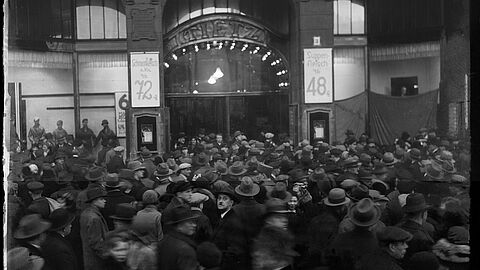 Das Bild zeigt eine Menschenmenge vor dem Warenhaus Tietz. Rechts und Links vom Eingang sind Angebotsschilder für Fleischwaren angebracht. In der Bildmitte schaut ein Man mit Fedora und Schnauzer in die Kamera.