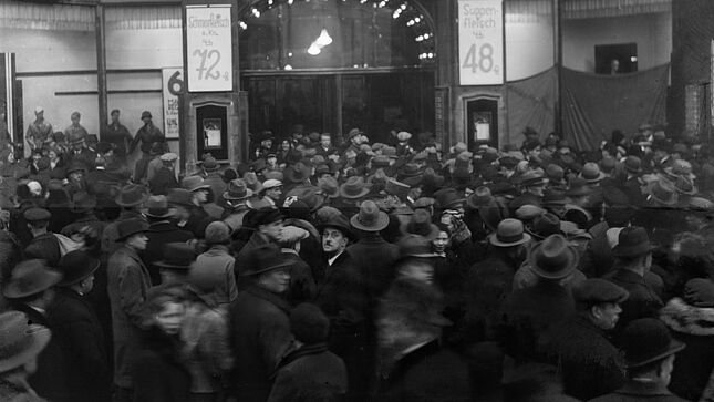 Das Bild zeigt eine Menschenmenge vor dem Warenhaus Tietz. Rechts und Links vom Eingang sind Angebotsschilder für Fleischwaren angebracht. In der Bildmitte schaut ein Man mit Fedora und Schnauzer in die Kamera.