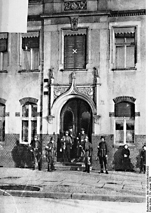 Eine Gruppe bewaffneter und mit Uniformen und Helmen bekleidete Männer steht vor einem Hauseingang. Rechts im Bild stehen zwei Kinder. Es handelt sich um eine Schwarz-Weiß-Aufnahme vom 15. Januar 1919.
