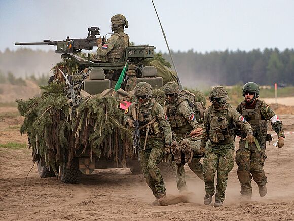 Vier Soldaten tragen einen weiteren Soldaten, der einen Verwundeten spielt, auf einer Bahre. An ihren Abzeichen erkennt man, dass drei Soldaten aus den Niederlanden stammen und einer aus Deutschland. Im Hintergrund ist ein getarntes Fahrzeug zu sehen, in dem ein Soldat am montierten Maschinengewehr sitzt.