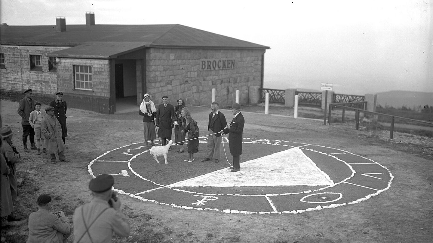 In einem mit unterschiedlichen Zeichen bemalten Hexenkreis stehen mehrere Leute sowie eine Ziege, die von einem der Männer an einer Leine geführt wird. Im Hintergrund sind Berge sowie ein Haus mit der Aufschrift „Brocken“ zu erkennen. Im Vordergrund stehen mehrere Männer mit Kameras, die von der Szene Fotos anfertigen., Quelle:
Bundesarchiv, Bild 102-13579 / Fotograf: Pahl, Georg