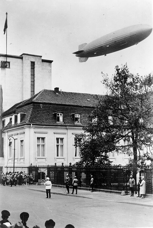 Einige Menschen stehen an einer Straße. Über einem Haus fliegt der Zeppelin LZ 129 „Hindenburg“. 