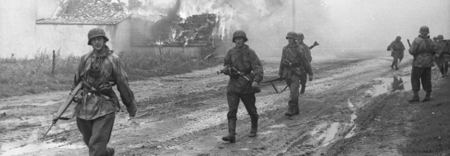 Deutsche Soldaten auf einer Straße vor brennenden Gebäuden.
