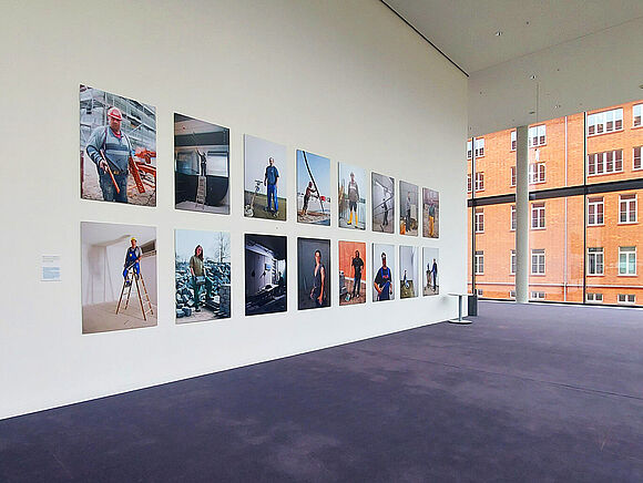 Zwei Reihen mit je fünf quadratischen farbigen glänzenden Bildern in einem Flur mit blauem Teppich und großer Fensterfront.