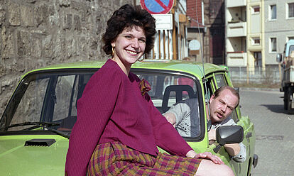 Frau präsentiert Modekollektion auf einem grünen PKW Wartburg sitzend, ein Mann schaut auf der Fahrerseite aus dem Auto