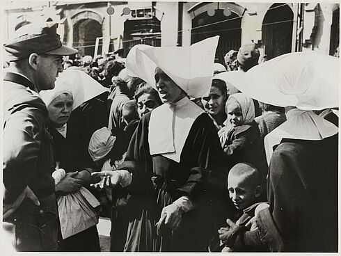 Eine polnische Schwester spricht mit einem deutschen Soldaten. Neben ihr stehen zwei weitere Schwestern, im Hintergrund mehrere Zivilistinnen und Zivilisten.