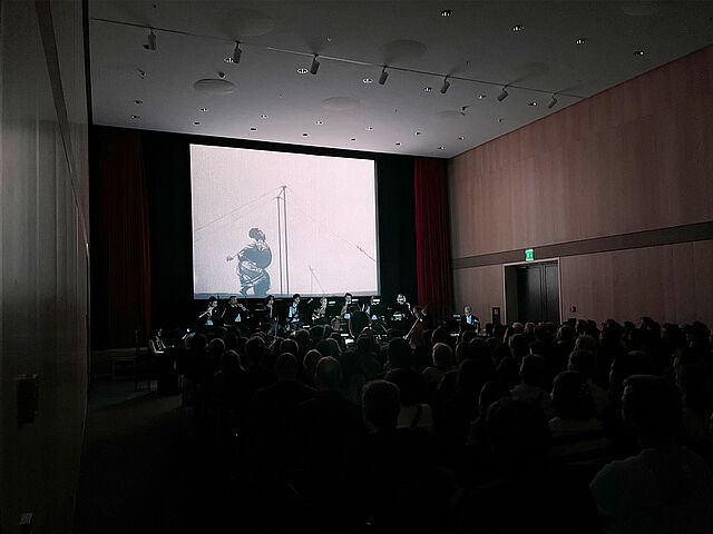 Ein dunkler Kinosaal mit sitzenden Menschen, die auf eine beleuchtete Leinwand schauen.