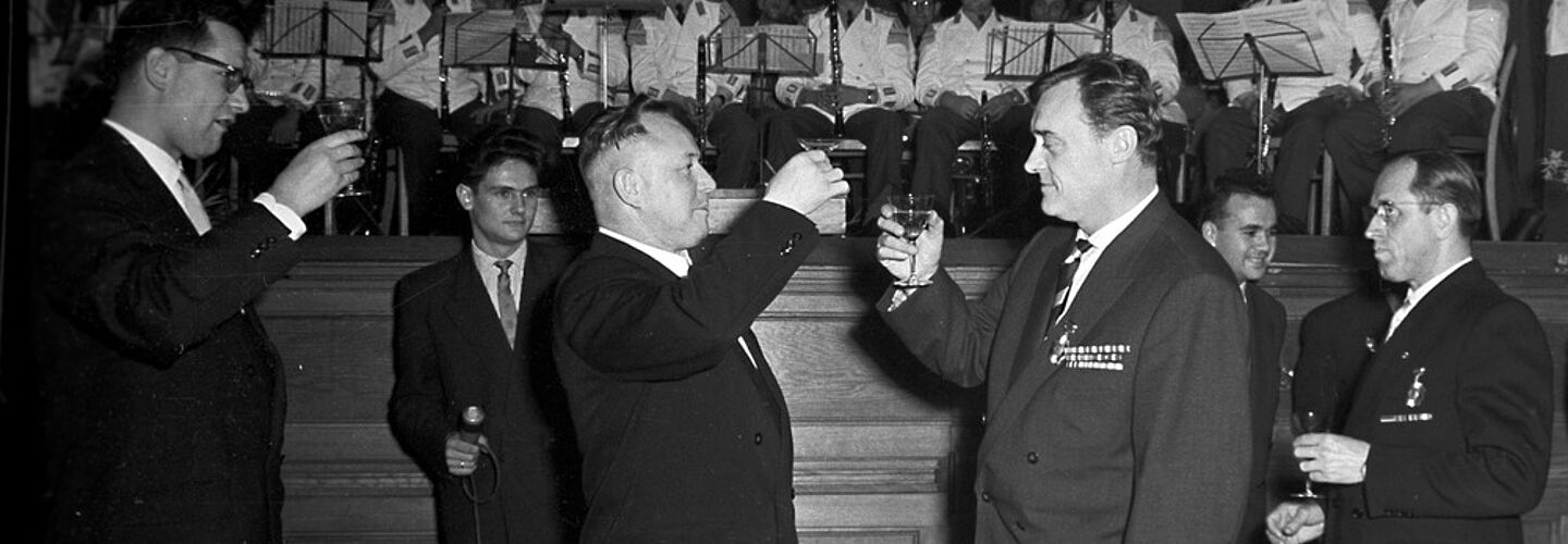 Schwarz-Weiß-Aufnahme eines Festbanketts in 'Haus 2' der Stasi-Zentrale in Berlin-Lichtenberg. Im unteren Bildabschnitt ist Tafel aufgebaut mit ahlreichen Gläsern und Flaschen zu erkennen. Hinter der Tafel stehen Männer in Anzügen. Etwa in der Bildmitte ist Stasi-Minister Erich Mielke zu sehen. Er prostet einem ihm gegenüberstehenden Mann zu. Die Männer stehen vor einer Bühne. Auf der Bühne befindet sich ein Orchester. Die Personen sitzen auf Stühlen. Sie haben zum Teil unterschiedliche Instrumente in der Hand. Zu erkennen sind zum Beispiel Klarinetten. Vor ihnen sind Notenständer aufgebaut. Darauf sind die Worte: 'Dank euch, den Söhnen der Sowjetunion' zu erkennen.