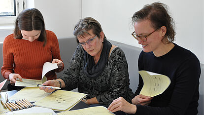 Drei Frauen an einem Tisch. Sie arbeiten mit auf dem Tisch liegenden kopierten Aktenseiten.