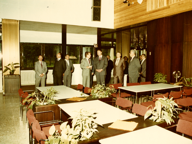Das farbige Lichtbild wurde durch eine nach außen geöffnete Tür aufgenommen. Von einigen Anwesenden werden Günther Müller und Gerhard Lange dabei beobachtet, wie sie zur Eröffnung ein rotes Band durchschneiden.