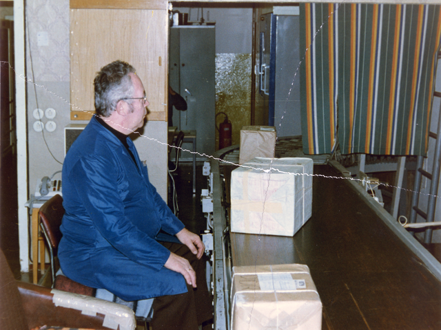 Das Bild zeigt einen Mann in einem blauen Arbeitskittel, der auf ein Förderband schaut, auf dem Pakete transportiert werden.