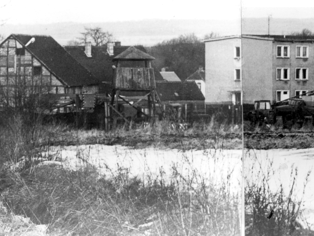[Das abgebildete schwarzweiße Lichtbild ist ein aus drei Fotos zusammengestelltes Panoramabild. Hinter schneebedeckter Freifläche ist abgezäuntes Gelände. Links steht ein Fachwerkgebäude, direkt rechts daneben ein hölzerner Aussichtsturm. In der Bildmitte ist ein dreistöckiges, langgestrecktes helles Gebäude mit Flachdach. Es folgt rechts daneben ein weiteres, etwas niedrigeres Gebäude mit Spitzdach, das aber hinter kahlen Laubbäumen schlechter erkennbar ist. Vor den Gebäuden sind, vermutlich militärische, Planenfahrzeuge aufgereiht.]