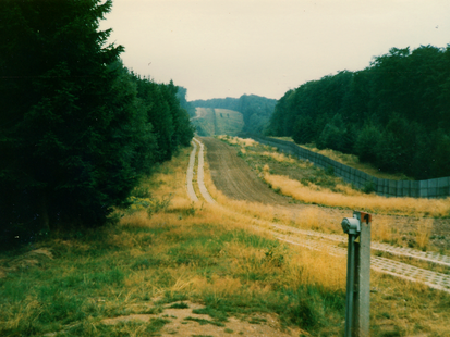 Zu sehen ist ein Blick entlang der Grenzsicherungsanlagen der DDR. Nach einem zweispurigen Weg aus Betongittern grenzt eine geharkte Sandfläche an. Vor dem Grenzzaun ist ein ungepflegt wirkender Abschnitt mit einigen hohen Gräsern. Auf der BRD-Seite grenzt Wald.