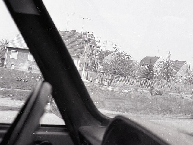 [Die schwarz-weiß Fotografie wurde durch die Windschutzscheibe eines Fahrzeugs von der Beifahrerseite über das Lenkrad hinweg aufgenommen. Auf der Fotografie sind im Hintergrund Einfamilienhäuser zu sehen. Links ist ein Einfamilienhaus mit Baugerüst zu erkennen. Rechts am Rand des Bildes ist ein parkendes Transportfahrzeug mit Beifahrer zu erkennen.]