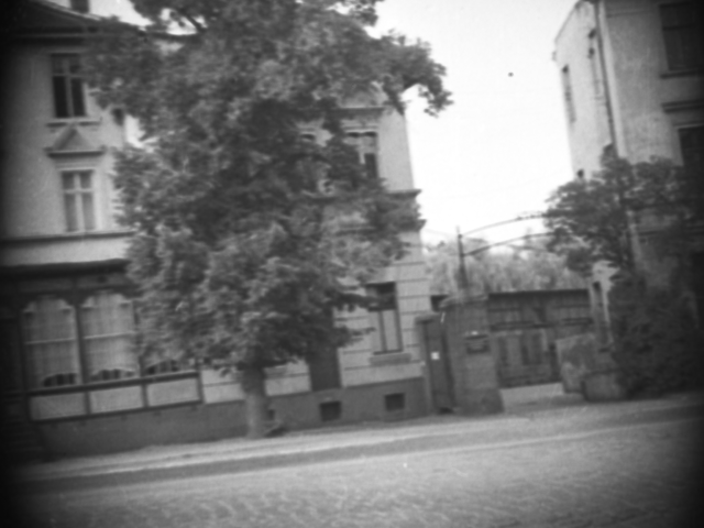 Vom gegenüberliegenden Gehweg aus wurde mit versteckter Kamera eine Toreinfahrt zwischen zwei Häusern am Rand einer unbekannten kopfsteingepflasterten Straße aufgenommen.