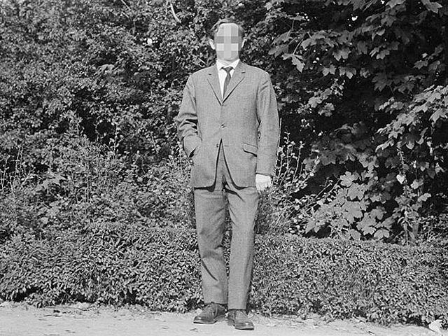 Es handelt sich um ein schwarz-weißes Lichtbild. Die Frontalaufnahme zeigt einen Mann im Anzug vor einer Hecke.