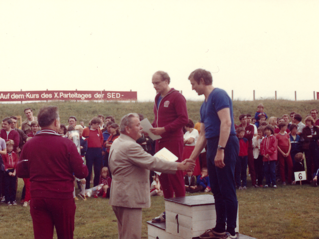 [Das farbige Lichtbild zeigt eine Siegerehrung bei einem sportlichen Wettkampf im Freien. Ein Mann im Anzug gratuliert dem Zweitplatzierten auf dem Treppchen mittels Handschlag und hält Urkunden bereit. Im Hintergrund stehen Zuschauer, darunter einige Sportler und Familien. Auf einer Anhöhe ist ein Banner gespannt.] Auf dem Kurs des X. Parteitages der SED.