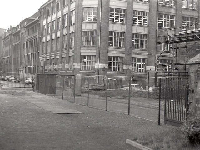 [Von einem eingezäunten, ebenmäßigen Gelände aus wurde eine Straßenecke fotografiert. Dabei stören mehrere Zäune das Blickfeld. Auf der anderen Straßenseite ist ein Altbau mit runder Ecke, auf beiden Straßenseiten parken Autos. Auf dem rechts angrenzenden Grundstück steht ein Gerüst an einem Backsteingebäude. Das Fotonegativ ist schwarzweiß.]