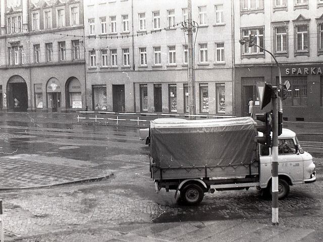 [Auf dem schwarzweißen Negativ wurde eine Straßenkreuzung aufgenommen. Im Vordergrund ist eine Verkehrsampel zu sehen, dahinter biegt nach rechts ein Lastkraftwagen mit Plane beim Rechtsabbiegen zu sehen. Es handelt sich um eine T-Kreuzung, auf der gegenüberliegenden Straßenseite ist eine Häuserzeile mit Geschäften zu sehen. Am markantesten ist der Schriftzug einer Sparkassen-Filiale.]