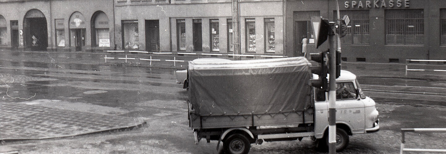 [Auf dem schwarzweißen Negativ wurde eine Straßenkreuzung aufgenommen. Im Vordergrund ist eine Verkehrsampel zu sehen, dahinter biegt nach rechts ein Lastkraftwagen mit Plane beim Rechtsabbiegen zu sehen. Es handelt sich um eine T-Kreuzung, auf der gegenüberliegenden Straßenseite ist eine Häuserzeile mit Geschäften zu sehen. Am markantesten ist der Schriftzug einer Sparkassen-Filiale.]