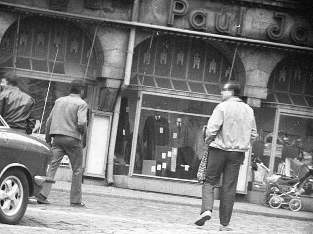 [Auf dem Schwarz-Weiß-Negativ wurde eine Schaufensterzeile eines alten Hauses mit schmuckreicher Fassade abgebildet. Von der Außenreklame ist nur "Paul Ja" zu lesen, in dem mittleren Schaufenster sind Schaufensterpuppen in Herrenanzügen zu sehen. Vier Passanten haben überwiegend den Rücken zur Kamera gewandt, am linken Bildrand ist das Heck eines Autos zu erkennen. Die Aufnahme wurde verdeck von einem gepflasterten Platz aus aufgenommen, erkennbar an dem Vignetteneindruck des dunklen Bildrandes.]