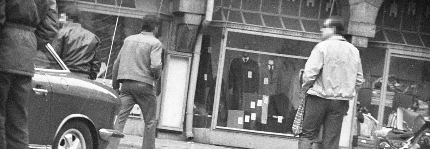 [Auf dem Schwarz-Weiß-Negativ wurde eine Schaufensterzeile eines alten Hauses mit schmuckreicher Fassade abgebildet. Von der Außenreklame ist nur "Paul Ja" zu lesen, in dem mittleren Schaufenster sind Schaufensterpuppen in Herrenanzügen zu sehen. Vier Passanten haben überwiegend den Rücken zur Kamera gewandt, am linken Bildrand ist das Heck eines Autos zu erkennen. Die Aufnahme wurde verdeck von einem gepflasterten Platz aus aufgenommen, erkennbar an dem Vignetteneindruck des dunklen Bildrandes.]