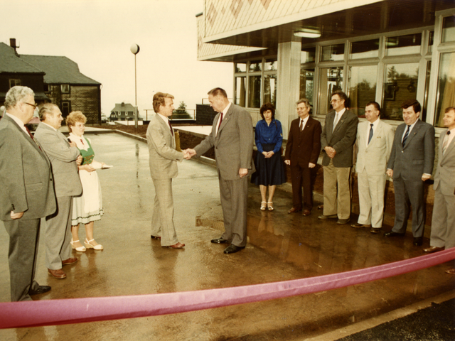 Umringt von mehreren Herren im Anzug sowie zwei Damen in Röcken schütteln sich im Mittelpunkt des Bildes zwei Herren, Günther Müller und Gerhard Lange, die Hand. Sie stehen vor einem luftig gestalteten Gebäudekomplex, der Boden ist betoniert. Den Bildvordergrund am unteren Rand bildet ein breites rotes Band. Es handelt es sich um ein farbiges Lichtbild.