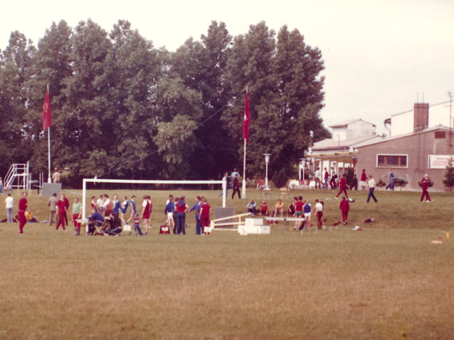 [Auf dem Fußballplatz findet Kugelstoßen statt. Viele Sportler stehen in kleinen Gesprächsgruppen zusammen oder sitzen auf dem Rasen. Hinter dem Tor stehen drei Fahnenmasten, zwei mit roten Fahnen, die dritte mit der Fahne der DDR gehisst. Rechts im Bildhintergrund des farbigen Lichtbilds ist eine Art Vereinshaus von der Schmalseite zu sehen, dessen Terasse überdacht ist. Links daneben grenzt eine Baumgruppe.]