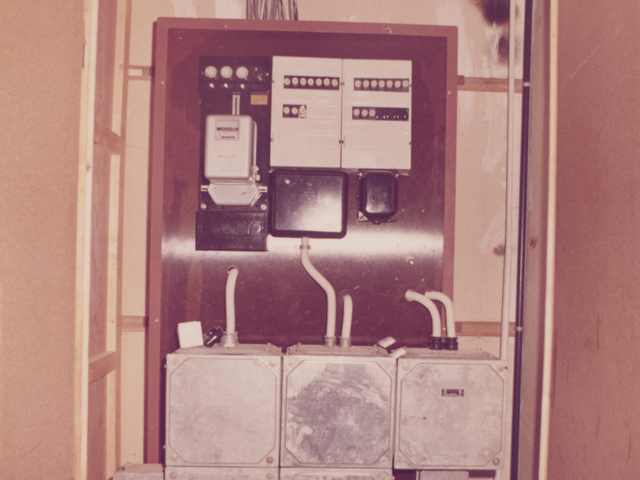 [Auf dem Farbfoto ist ein Stromzähler- und Sicherungskasten zu sehen. Die gesamte Geräteeinheit ist mit einem Holzgehäuse verkleidet, dessen geöffnete Tür die Sicht auf die Elemente freigibt. Die einzelnen Geräte und Elemente sind an eine braune Holzrückwand montiert, in der sich gebohrte Löcher befinden, durch die die Kabel weiterführen.]