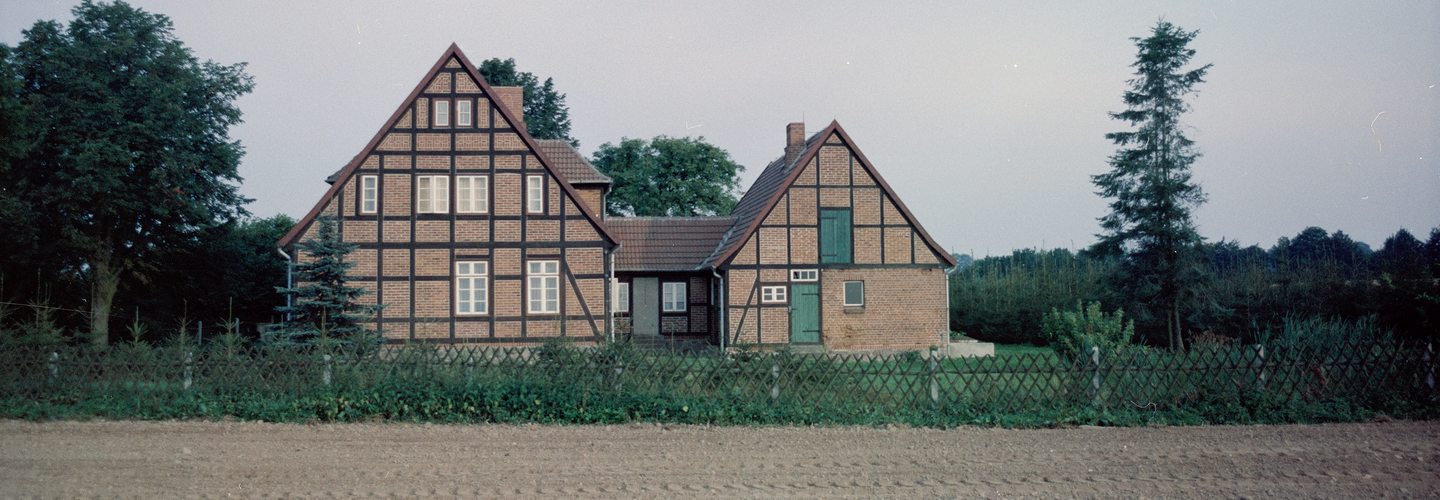 Das Farbfoto zeigt einen Fachwerkhof mit drei Seiten am rande eines Ackers. Das Grundstück des Hofes ist mit einem Zaun begrenzt. Auf dem Grundstück und daneben wächste grüne Vegetation.