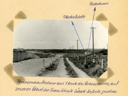 [Das schwarzweiße Lichtbild zeigt einen Blick in die Grenzsicherungsanlagen der DDR. Von links nach rechts sind zu sehen: der Metallzaun, daran angrenzend die geharkte Sandfläche. Ein unbefestigter, aber ausgefahrener Weg wird auf beiden Seiten mit Betonpfeilern markiert. Weiter rechts davon befindet sich ein Grünstreifen, auf dem Elektromasten entlang der Grenzlinie aufgestellt sind. Im Hintergrund liegt eine Kontrollstelle für die Transitstraße. Darum herum sind verstärkt Laternen aufgestellt, ein Wachturm ist in der Nähe. Zwei Stellen wurden auf dem Foto mit blauem Kugelschreiber mittels Pfeilen markiert, die außerhalb des Bildes wie folgt beschriftet wurden:] 1. Kontrollstelle Postenturm [handschriftlich mit blauem Kugelschreiber: Panoramaaufnahme vom 1.Knick des Grenzzaunes, auf unseren Gebiet zur Transitstraße Lübeck-Saßnitz gesehen]