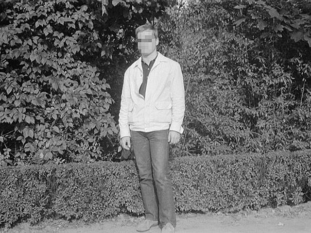 Die Frontalaufnahme zeigt einen Mann in heller Jacke und dunkler Hose vor einer Hecke. Es handelt sich um ein schwarz-weißes Lichtbild.