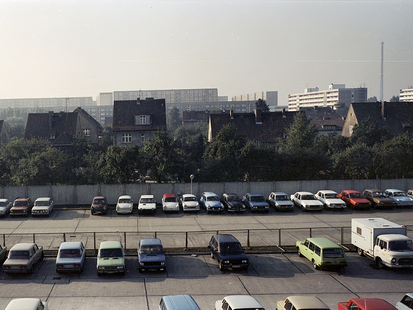 Eine farbige Weitwinkelaufnahme zeigt einen Betonparkplatz vor Einfamilienhäusern im Grünen, im Bildhintergrund sind Hochhäuerreihen zu erkennen. Auf dem Parkplatz sind ordentlich senkrecht geparkte Reihen von verschiedenen Pkw-Modellen und in verschiedensten Farben zu sehen. Dazwischen gibt es ofetmals Lücken, die auch deutlich größer Ausfallen können.