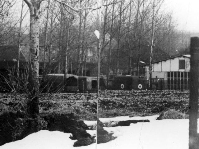 [Zu sehen ist ein scharzweißes Lichtbild, das aus zwei horizontal aneinander geklebten Fotos besteht. Hinter einem schmalen Streifen eines geegtes Feldes grenzt ein Zaun. Auf dem Gelände sind diverse, vermutlich militärische, Planenfahrzeuge geparkt, darunter ein vermuteter Sanitätswagen, der mit Kreuz auf weißem Kreuz gekennzeichnet ist. Die Fahrzeuge stehen vor einem hellen Flachbau mit großem, geschlossenem Tor. Weitere mehrgeschossige Häuser und diverse kahle Laubbäume sind im Hintergrund. Es liegt Schnee.]