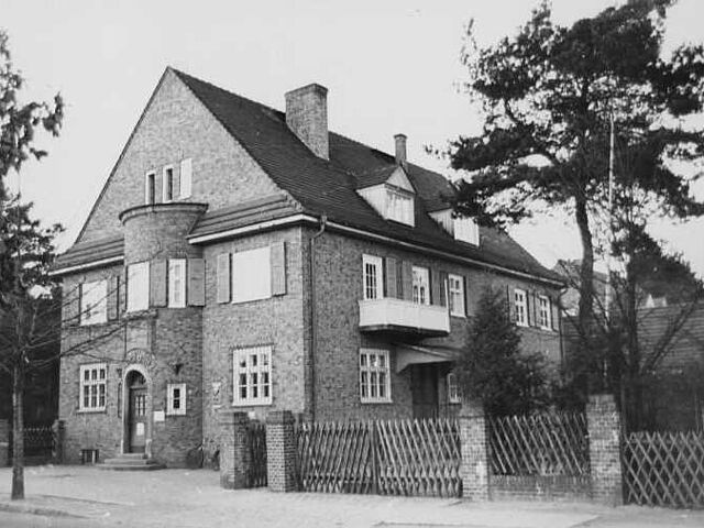 Zu sehen ist ein Backsteingebäude, zwei Etagen mit Spitzdach und Erker mit Eingangstür an der Schmalseite. Hinter einem Jägerzaun rechts am Haus liegt ein weitere EIngang und einige Nadelhölzer.