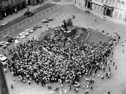 Das Schwarzweißfoto zeigt einen Blick auf den Altstädter Ring in Prag in nordwestlicher Richtung. Das Bild ist von oben aufgenommen, vermutlich vom Turm des altstädtischen Rathauses aus. In der Mitte des Platzes ist das Jan-Hus-Denkmal zu sehen. Es ist zum Teil mit Transparenten behängt, einzelne Menschen sitzen auf dem Sockel. Davor, in Richtung Süden, hat sich eine große Menschentraube versammelt, die in Richtung des Denkmals blickt. Vermutlich hält dort jemand eine Rede.