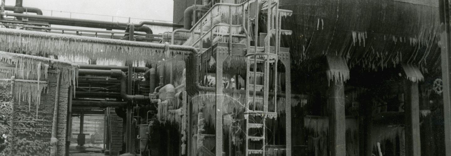 [Auf der schwarz-weiß Fotografie ist ein Teil eines Gebäudes auf einem Industriegebiet zu sehen. An dem Gebäude entlang verlaufen einige Rohre. Unter den Rohren ist ein Durchgang zu erkennen. Rechts vor dem Gebäude ist ein waagerechter Tank angebracht. Zum Tank hinauf führt eine Leiter mit Schutzvorrichtung. Unten rechts auf der Fotografie ist ein Seil vermutlich zur Abtrennung des Bereiches angebracht worden. Daran sind einige Hinweiswimpel mit Ausrufezeichen befestigt. Das Foto ist im Winter aufgenommen worden, da auf dem Boden Schnee zu erkennen ist und der Tank, die Leiter und die Rohre mit vielen Eiszapfen überzogen sind.]