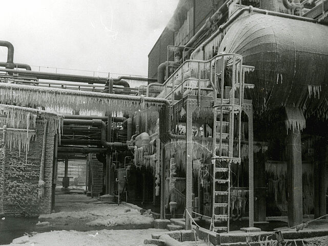 [Auf der schwarz-weiß Fotografie ist ein Teil eines Gebäudes auf einem Industriegebiet zu sehen. An dem Gebäude entlang verlaufen einige Rohre. Unter den Rohren ist ein Durchgang zu erkennen. Rechts vor dem Gebäude ist ein waagerechter Tank angebracht. Zum Tank hinauf führt eine Leiter mit Schutzvorrichtung. Unten rechts auf der Fotografie ist ein Seil vermutlich zur Abtrennung des Bereiches angebracht worden. Daran sind einige Hinweiswimpel mit Ausrufezeichen befestigt. Das Foto ist im Winter aufgenommen worden, da auf dem Boden Schnee zu erkennen ist und der Tank, die Leiter und die Rohre mit vielen Eiszapfen überzogen sind.]